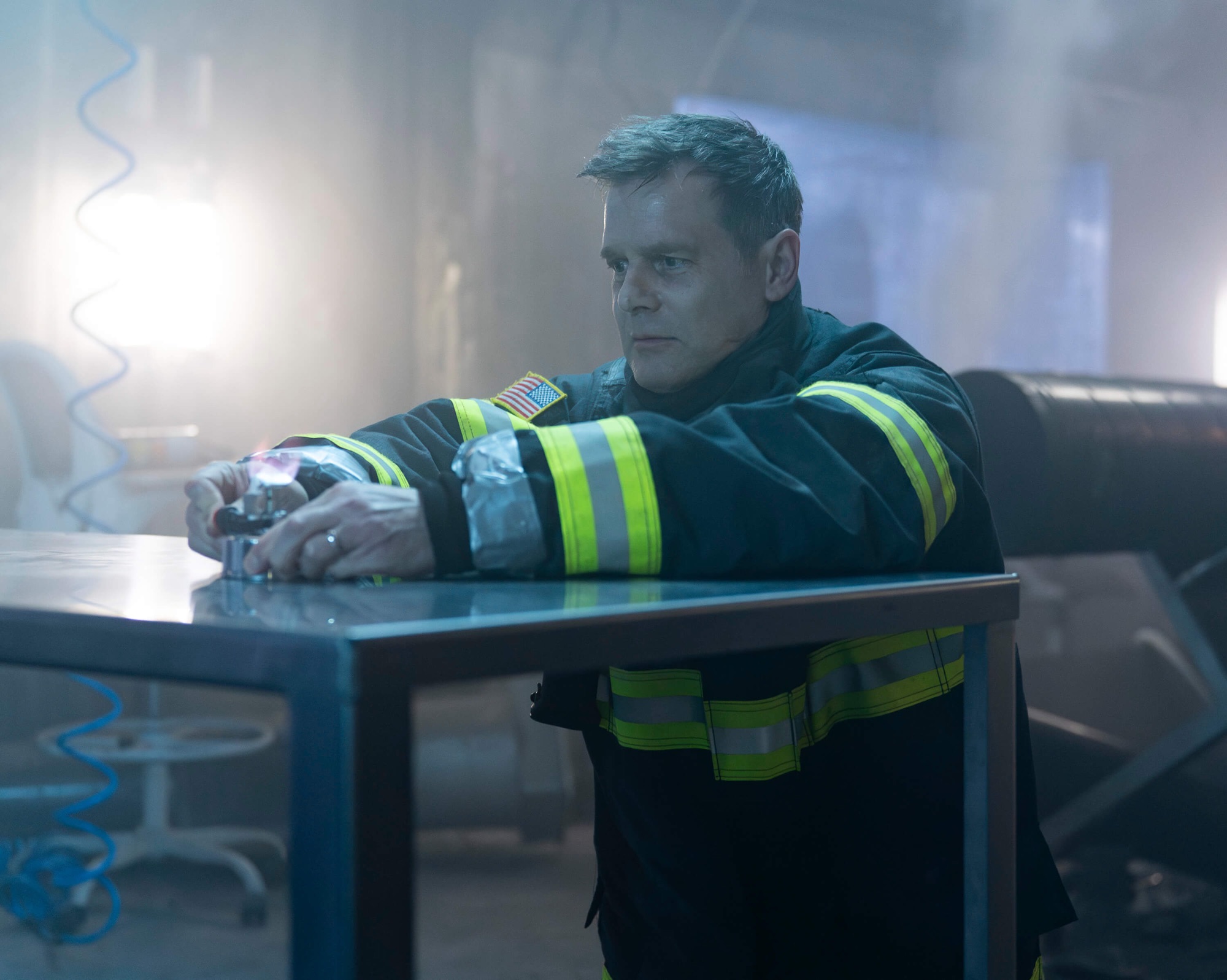 A firefighter in a black uniform with yellow reflective stripes crouches behind a metal table, gripping a small device emitting a faint flame. He looks intensely focused, with mist and industrial equipment in the background, and dramatic lighting casting shadows around him.